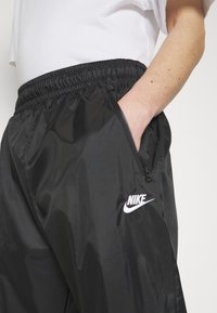 Černé, lehké joggingové kalhoty s elastickým pasem a kapsou na zip. Na levé noze se nachází vyšívané logo Nike v bílé barvě. Hladká textura.