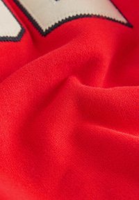 Gros plan sur un tissu rouge, texture douce, finition lisse. Couture distinctive visible le long des bords, mettant en avant un design sportif avec une couleur vive.