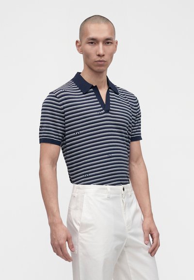 Homme debout portant un polo à manches courtes rayé bleu marine et blanc et un pantalon blanc taille haute sur un fond uni.