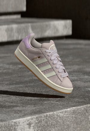 Jasnofioletowy sneaker Adidas Campus z białymi paskami i gumową podeszwą, unoszący się nad teksturowanymi betonowymi schodami.