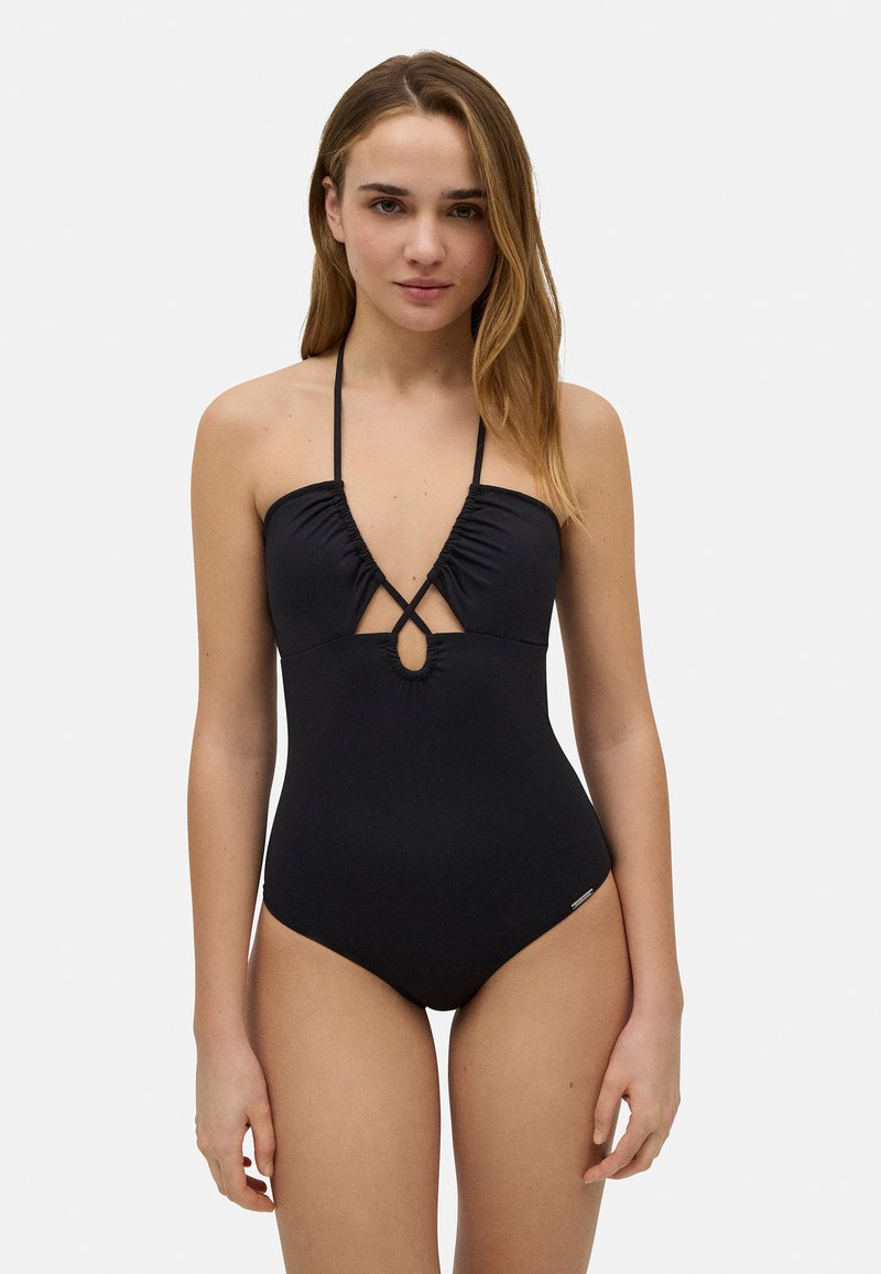 Bañador negro de una pieza con fruncido en el busto, escote en V profundo y tirantes cruzados en la espalda. Presenta un corte en forma de llave en el centro. Textura de tela suave.