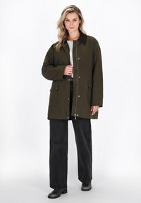 Femeie îmbrăcată într-o haină închisă la culoare cu buton și fermoar de culoare verde închis, bluză albă, pantaloni largi negri și cizme negre, stând pe un fundal alb.