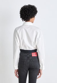 Camicia bianca oversize con colletto alto, abbinata a jeans neri a vita alta con un elastico in vita di colore rosso. Tessuto dalla superficie liscia.