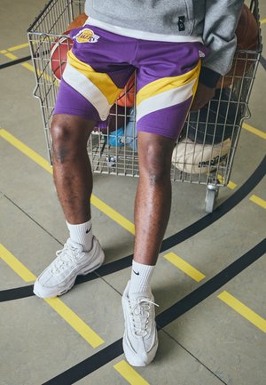 Lilla og gule basketballshorts med chevron-mønster, parret med hvide sneakers og sokker. Basketballer synlige i en metalvogn.