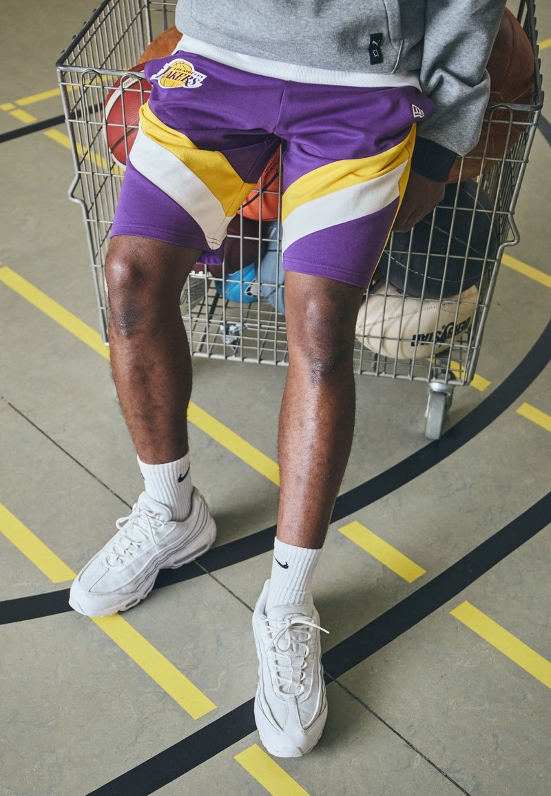 Pantalones cortos de baloncesto morados y amarillos con un patrón de chevron, combinados con zapatillas y calcetines blancos. Balones de baloncesto visibles en un carro de metal.