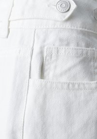 Witte denimstof met een verhoogde textuur, voorzien van een klein voorvak en metalen knoopversieringen. De stiksels zijn zichtbaar langs de randen.
