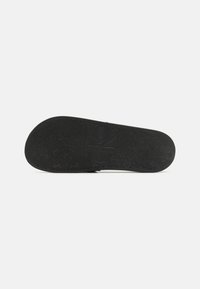 Semelle en caoutchouc noir avec un motif texturé et un logo en relief. Design plat avec des bords arrondis pour le confort et la stabilité.