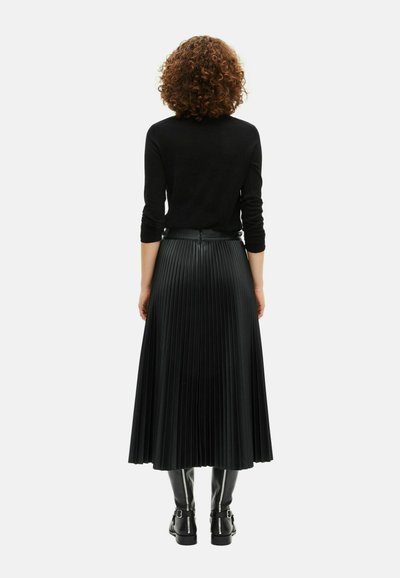 Γυναίκα με σγουρά μαλλιά, φορώντας μαύρο μπλούζα με μακριά μανίκια, πλισέ μαύρη midi φούστα και μαύρες μπότες μέχρι το γόνατο με αγκράφες, όρθια και στραμμένη με πλάτη.