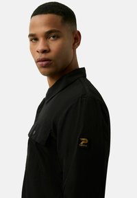 Zwarte button-up shirt met een kraag, voorzien van een borstzak en een gouden logo patch op de mouw. Structureerde stof en een getailleerde pasvorm.
