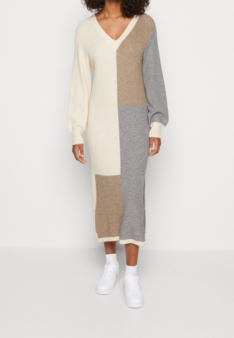 Femme portant une robe longue en tricot à blocs de couleur crème, beige et gris, avec des manches bouffantes et des baskets blanches, debout devant un fond uni.