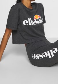 Grå kortärmad t-shirt med "ellesse"-logotyp i vitt, parat med matchande grå byxor med upprepad vit text. Blandning av bomull, avslappnad passform.