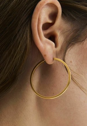 Nahaufnahme eines Ohrs mit glatter Haut und hellbraunem Haar, das einen großen dünnen goldenen Creolen-Ohrring trägt.