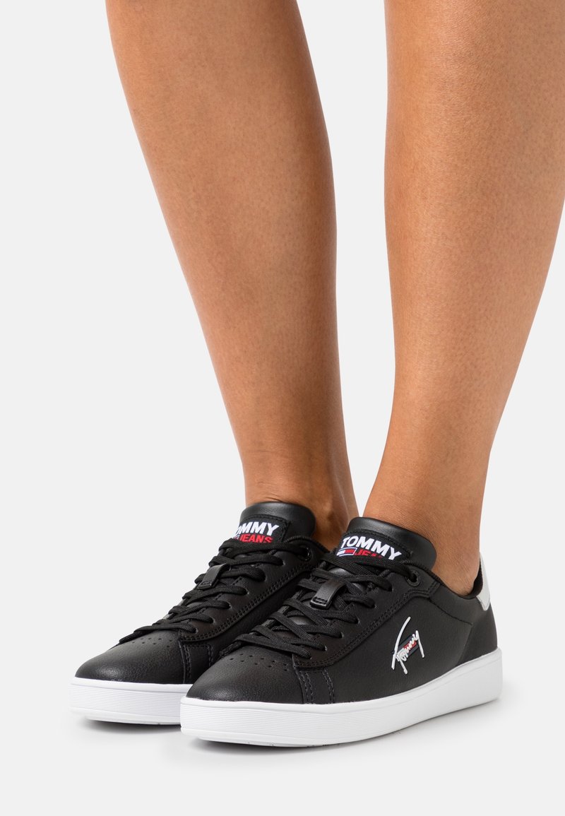 Personne portant des baskets basses en cuir noir avec des semelles blanches et des logos Tommy Jeans sur la languette et le côté, debout sur un fond uni.