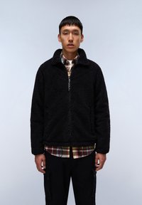 Giacca in fleece nera con collo alto, zip frontale e tasche laterali, indossata sopra una camicia a quadri multicolore, esposta su uno sfondo chiaro.