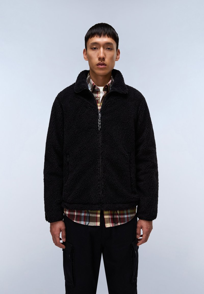 Giacca in fleece nera con collo alto, zip frontale e tasche laterali, indossata sopra una camicia a quadri multicolore, esposta su uno sfondo chiaro.