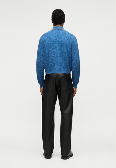 Άτομο που στέκεται με την πλάτη στραμμένη προς εμάς, φοράει μπλε πουλόβερ με μακριά μανίκια και μαύρο δερμάτινο παντελόνι μπροστά σε απλό ανοιχτό φόντο.
