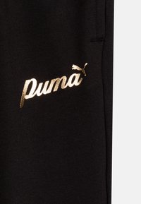 Melnas sporta krekla audums ar lielu, spīdīgu zeltītu "Puma" logo un stilizētu kaķa simbolu, kas izvietots izteiksmīgi kreisajā pusē.