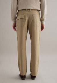 Persona con pantalones beige de pierna recta con solapa y bolsillos traseros de parches, combinados con zapatos marrones y un suéter beige, de pie sobre un suelo gris.