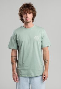 Vaaleanvihreä puuvillainen t-paita, jossa on lyhyet hihat, pyöreä kaulus ja painettu logo vasemmassa rintapielessä. Mallilla on päällään väljät farkut.