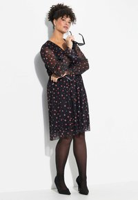 Schwarzes Kleid mit durchsichtigen langen Ärmeln, versehen mit einem Herzmuster in Rot und Rosa. Begleitet von schwarzen Strumpfhosen und spitzen Absatzschuhen.