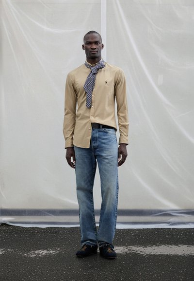 Μπεζ πουκάμισο με κουμπιά, μπλε φουλάρι με σχέδια, ανοιχτόχρωμο μπλε τζιν και σκούρα παπούτσια. Το μοντέλο στέκεται μπροστά σε ένα υφασμένο λευκό φόντο.