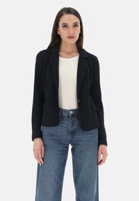 Blazer nero con revers, chiusura a un bottone e tasche frontali, abbinato a una maglietta bianca e jeans blu a vita alta. Texture liscia.