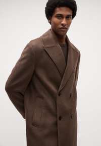 Cappotto doppio petto in lana marrone con rever a punta, bottoni neri e taschino sul petto. Il tessuto ha una texture morbida e una vestibilità su misura.