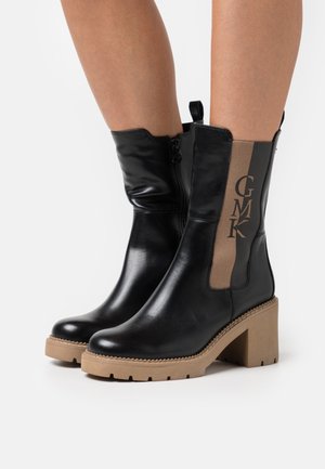Bottines en cuir noir avec un panneau latéral beige, dotées d'un talon épais et d'un logo en relief. Fermeture éclair et semelle texturée.