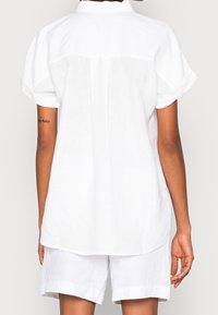 Een persoon in een losse witte korte mouw shirt en bijpassende witte broek, gezien vanaf de achterkant tegen een effen achtergrond.