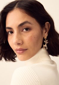 Gold-tone earrings featuring a reindeer design with sparkling accents and a snowflake detail. Model wears a ribbed white turtleneck.
