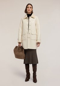 Cappotto bianco doppiopetto con colletto in shearling, due tasche frontali, indossato sopra un vestito a righe nere e beige, abbinato a stivali marroni con motivo a serpente.