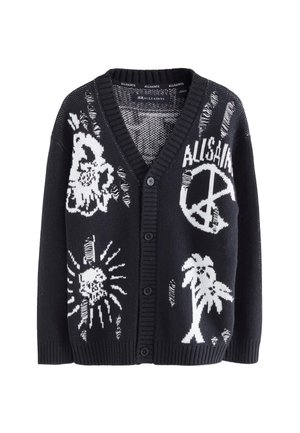 Jugendlicher Junge trägt eine schwarze Strickjacke mit weißen grafischen Designs über einem weißen T-Shirt und dunklen Jeans und steht vor einem grauen Hintergrund.