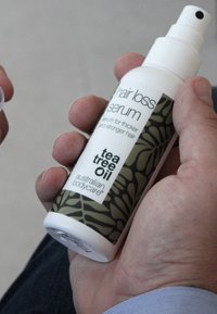 Håravfallsserum i en vit plastsprayflaska. Grön etikett med bladmönster. Texten inkluderar "teaträdolja" och "serum för tjockare hår."