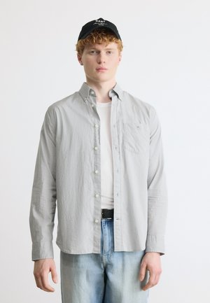 Jeune homme aux cheveux roux bouclés portant une casquette noire, une chemise boutonnée gris clair sur un t-shirt blanc, et un jean bleu clair sur fond blanc.