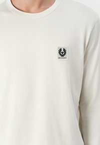 Long-sleeve, cream-colored t-shirt made of soft fabric featuring a small black and gray logo patch on the left chest area.