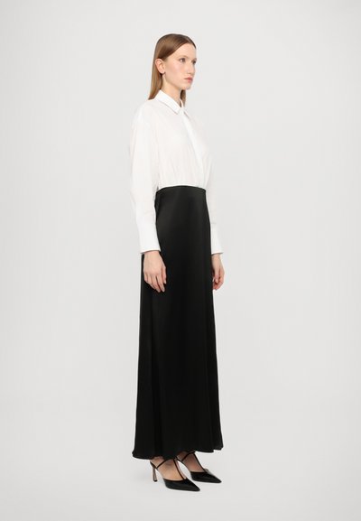 Lång svart klänning med satinliknande textur som har en vit krage och åtsittande svart midja. Modellen är matchad med svarta spetsiga klackar.