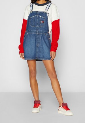 Kvinde iført en blå denim-overal-kjole med mærkede stropper over en hvid og rød langærmet sweater, kombineret med hvide sneakers med røde snørebånd.