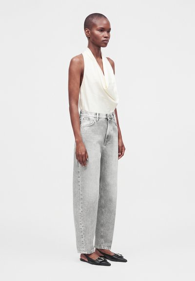 Femme aux cheveux courts portant un top blanc drapé sans manches, un jean gris clair taille haute et des ballerines noires pointues avec un détail boucle.