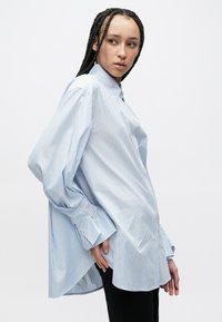 Niebiesko-biała koszula w paski z guzikami, z długimi rękawami, luźnym krojem, zaokrąglonym dołem i widocznym zapięciem na guziki. Delikatna faktura.