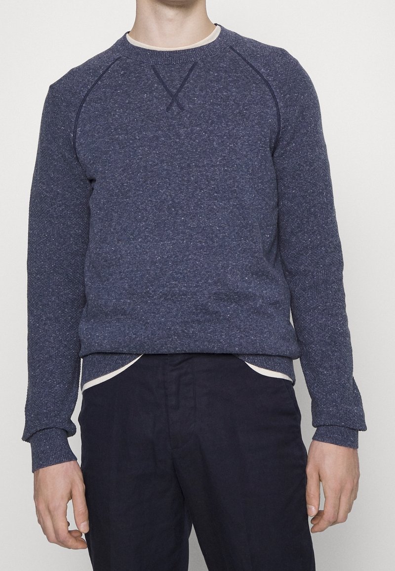 Homme portant un pull bleu à manches longues avec col rond et un pantalon sombre sur un fond clair uni.