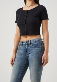 Camiseta negra corta con manga corta y cierre de botones en el frente. Presenta un sutil detalle de logo. Combinada con unos jeans ajustados de color azul claro.
