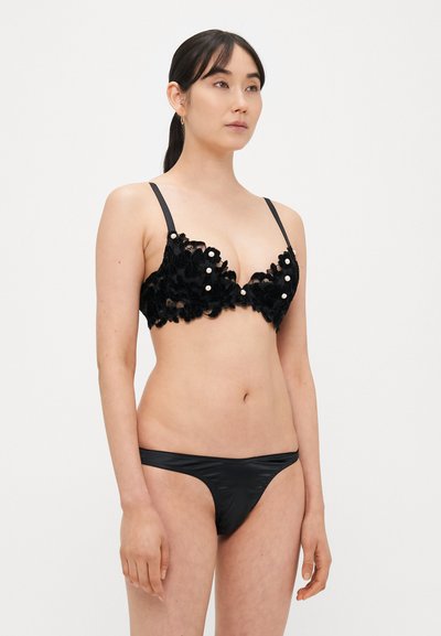 Črni čipkasti bralette z bisernimi poudarki in tankimi črnimi naramnicami, kombiniran s gladkimi črnimi tangicami. Oba zagotavljata oprijeto silhueto.