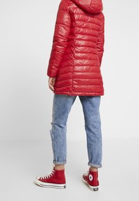 Abrigo acolchado rojo con capucha, diseño acolchado y acabado brillante. Combinado con jeans desgastados de color azul claro y zapatillas rojas de caña alta.