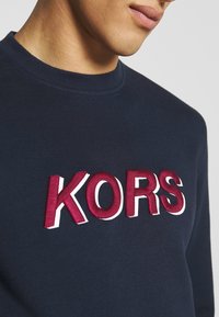 Námořnická modrá mikina se žebrovaným výstřihem, s výrazným vyšitým logem "KORS" v barvách bordó a bílá. Hladká textura.