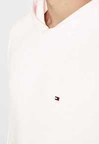 Sudadera rosa claro con cordón, que presenta un pequeño logotipo con acentos en rojo, blanco y azul en el pecho. Textura de tela suave.