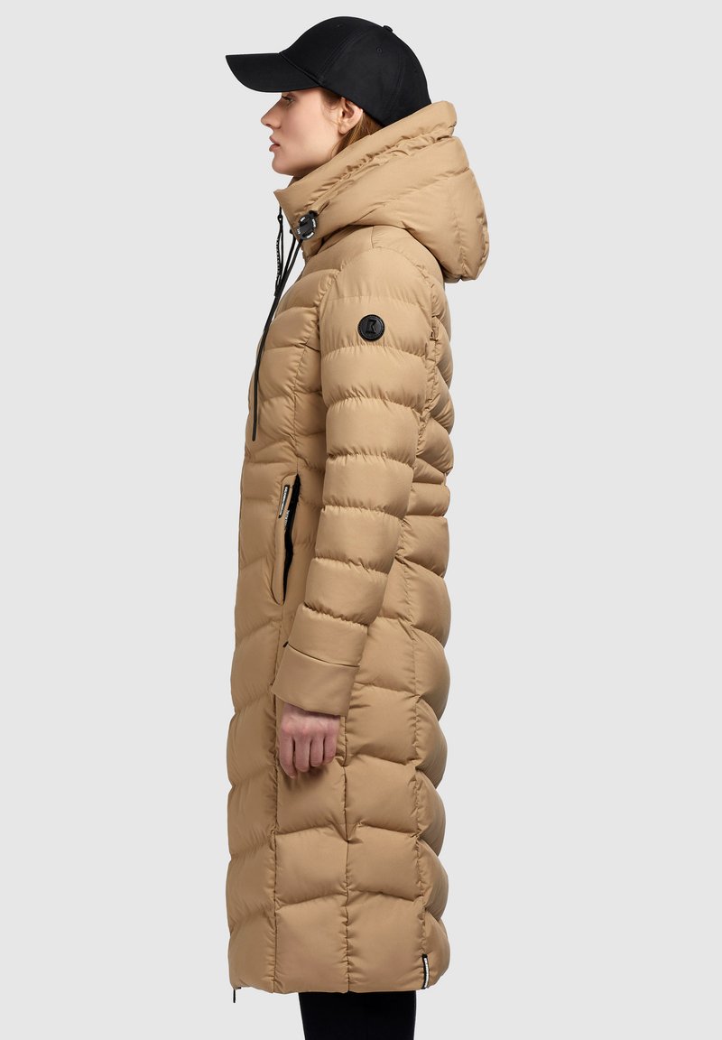 Beige gepolsterter langer Mantel mit hohem Kragen und Kapuze, mit seitlichen Reißverschlüssen und einem runden schwarzen Logo auf dem Oberarm, glatte Textur.