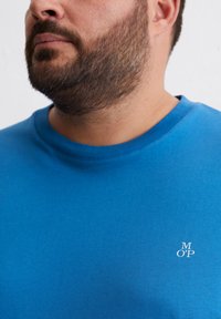Blauwe katoenen T-shirt met een ronde hals, korte mouwen en een klein wit logo op de linkerkant van de borst. Zachte textuur.