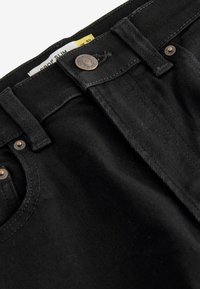 Jeans de mezclilla negra con corte ajustado. Cuenta con herrajes metálicos, diseño de cinco bolsillos y una etiqueta visible en el interior de la cintura.