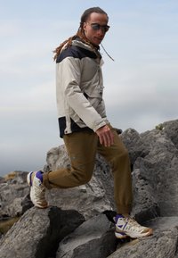 Utendørsjakke i lysegrå og svart, olivenbukser og beige tursko med lilla aksenter. Modell som trår på steinete terreng.