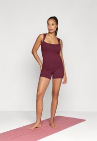 Μπορντό αθλητική αμάνικη μπλούζα με φαρδιές τιράντες και αντίστοιχα σορτς, κατασκευασμένα από μαλακό ύφασμα, πάνω σε ροζ στρωματάκι γιόγκα σε ελαφρύ φόντο.